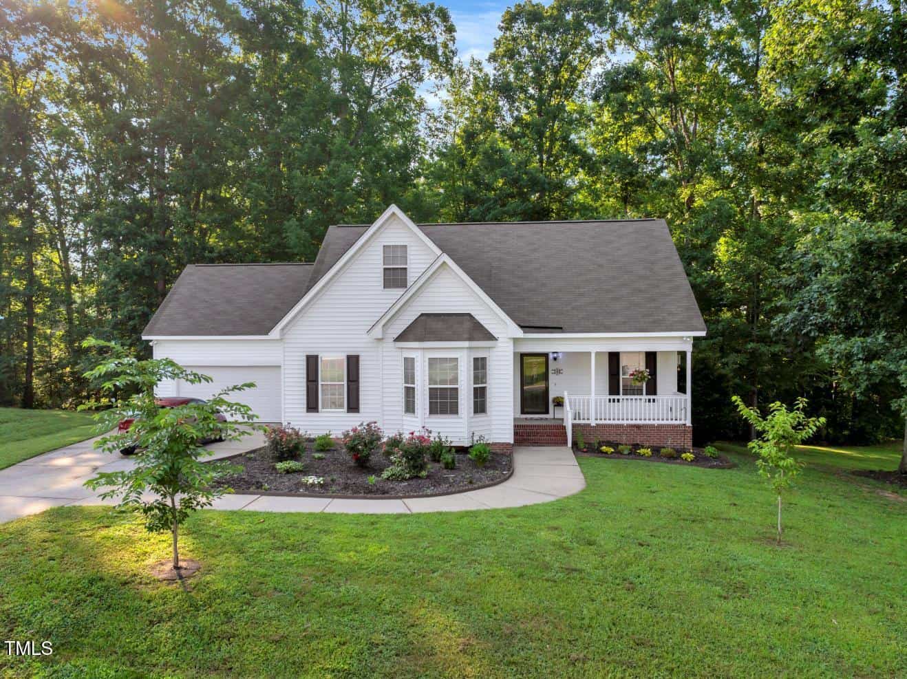 A white, single-story house with a gabled roof, front porch, and attached garage sits amid green grass and trees. A curved driveway and landscaped flower beds lead to the entrance. The yard is surrounded by forest.