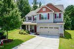 Two-story suburban house with gray siding, red-brown accents, and shutters. Attached two-car garage, covered front porch, landscaped lawn, trees, and a fire hydrant by the driveway. Surrounded by greenery and woods.