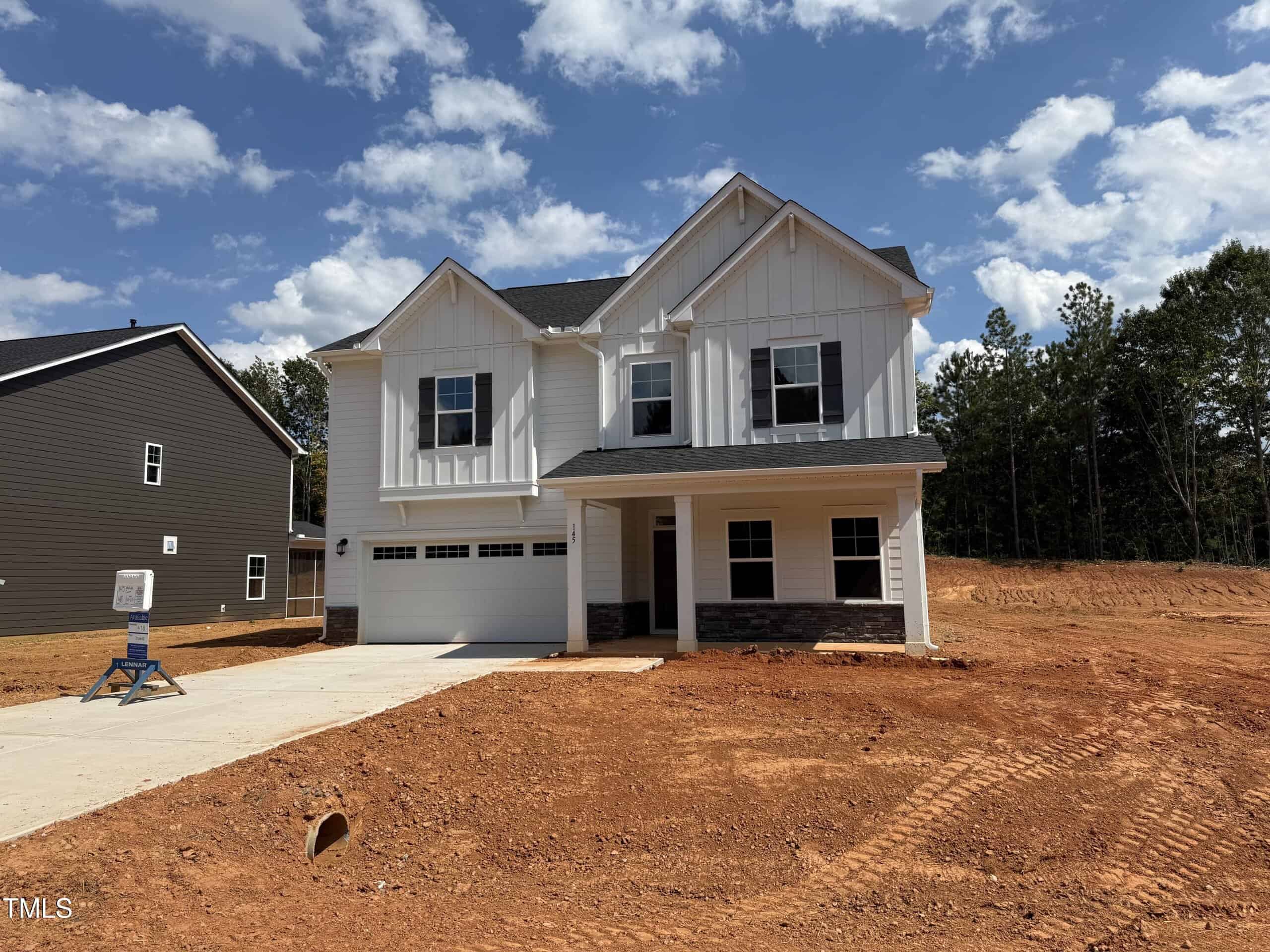 A newly constructed two-story white house with a covered front porch and attached garage sits on a lot with reddish-brown bare soil, surrounded by trees and neighboring homes under a blue sky with scattered clouds.
