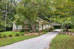 A light green house with a gabled roof and attached carport sits behind leafy trees and a white gravel driveway, surrounded by green grass, shrubs, and rocks on a summer day.