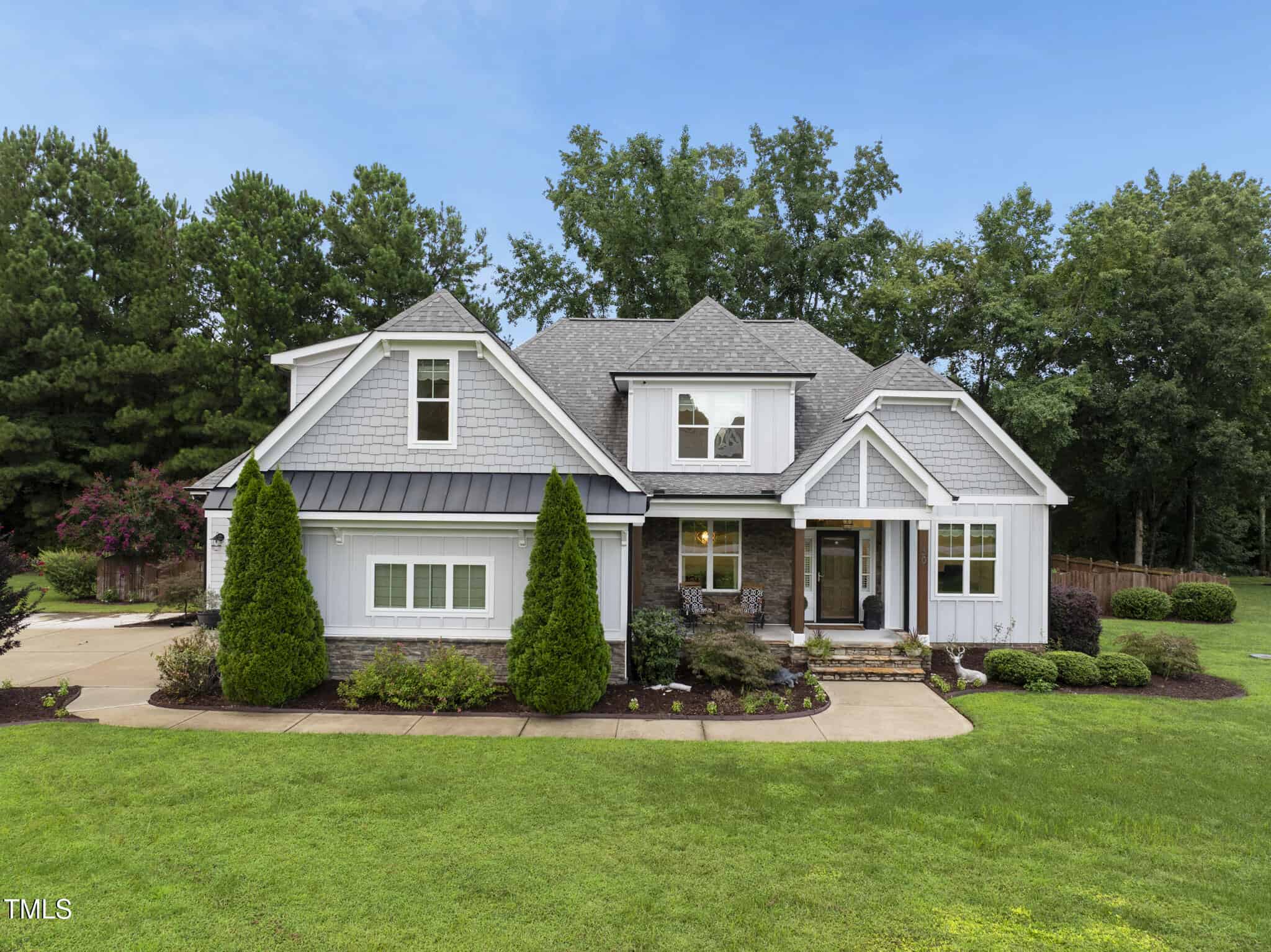 Modern two-story house with gray siding, white trim, and a black metal roof over the front porch, surrounded by green grass, landscaped bushes, and trees in the background under a clear blue sky.