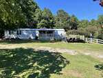 A white manufactured home with a covered front porch sits in a grassy yard surrounded by trees. A metal carport and white wooden fence are on the right, under a clear blue sky.