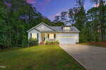 A single-story house with a front porch, white siding, and a two-car garage sits on a large, sloped lawn, surrounded by trees at dusk with a colorful sky in the background. The house and driveway are well lit.