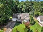 Aerial view of a two-story suburban house with a gray roof, white siding, and attached garage. The home is surrounded by green lawn, trees, and a driveway, with a detached building on the right.