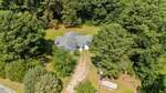 Aerial view of a blue house with a gray roof surrounded by tall trees and greenery. A dirt driveway leads to the house, with a covered boat parked beside it. Dense forest is visible in the background.