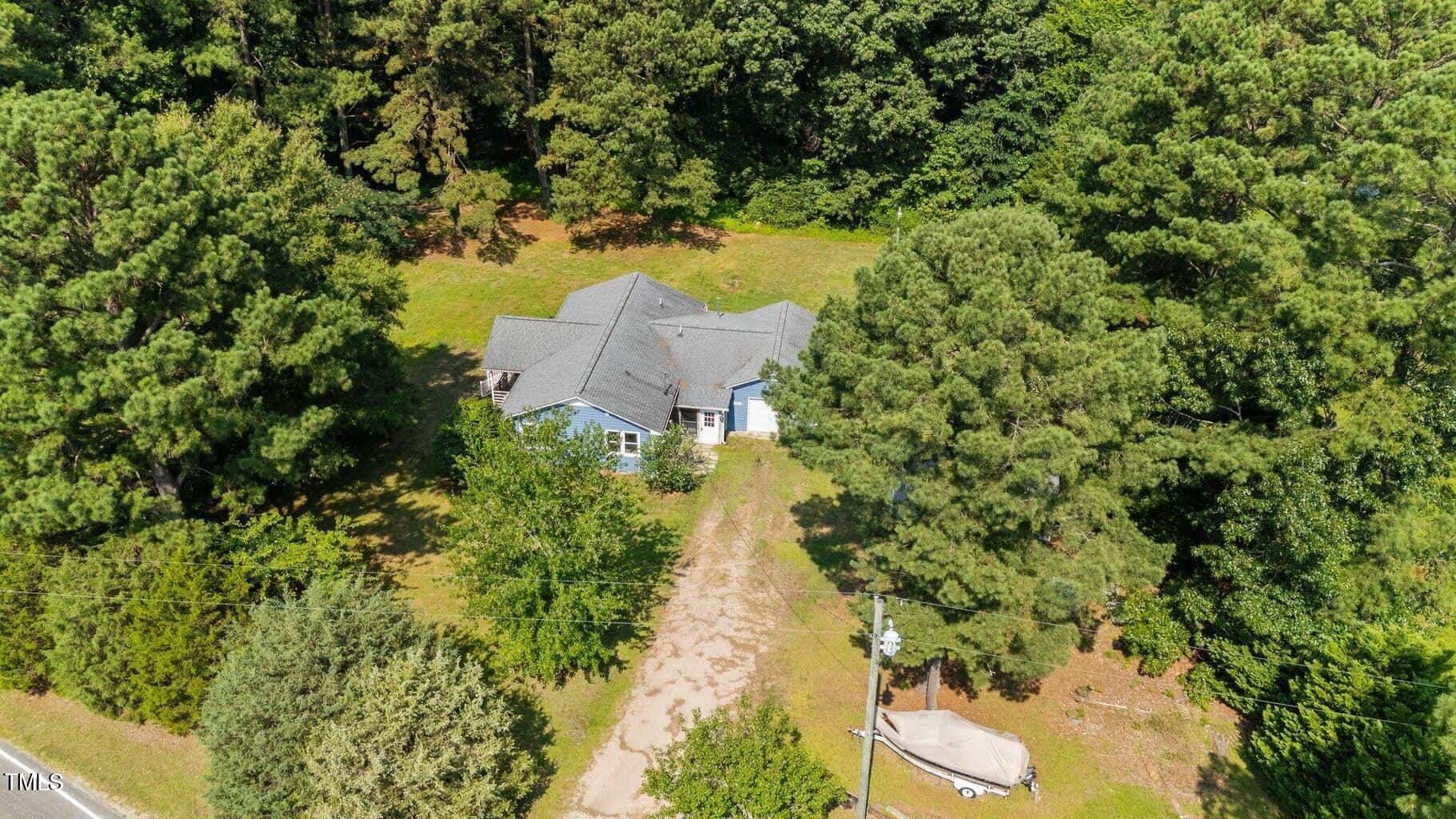 Aerial view of a blue house with a gray roof surrounded by tall trees and greenery. A dirt driveway leads to the house, with a covered boat parked beside it. Dense forest is visible in the background.