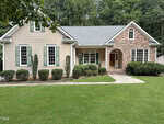 Single-story house with beige siding, green shutters, and a front entrance accented by stonework. The yard is well-maintained with shrubs and a lush green lawn, bordered by trees in the background.