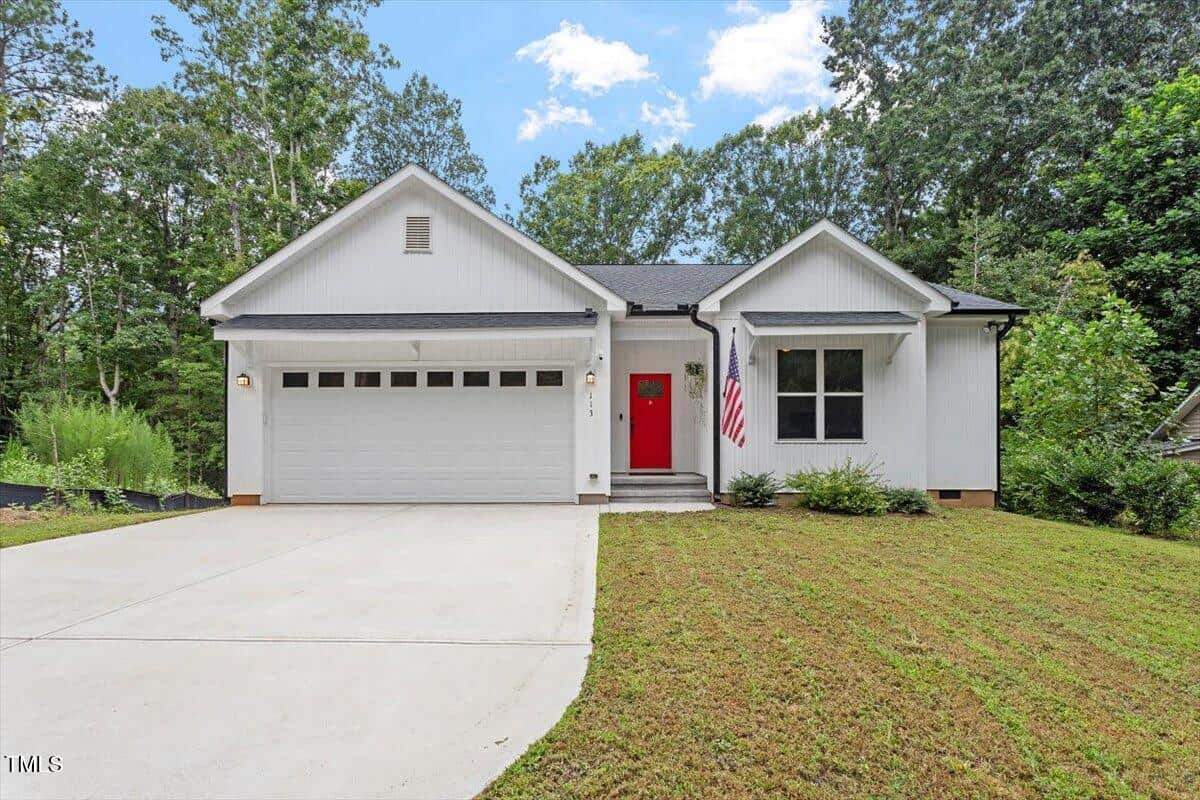 A modern white single-story house with a double garage, red front door, and American flag by the entrance. The home has a well-kept lawn and is surrounded by tall trees under a partly cloudy sky.