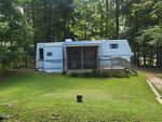 A large white RV with blue accents is parked in a wooded area, featuring an attached covered porch with a ramp leading to a grassy yard surrounded by tall trees.