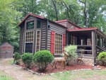 A cozy log cabin with red-trimmed windows, a covered porch, and two deer statues in the yard, surrounded by green bushes and tall trees, with a small shed in the background.