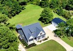 Aerial view of a two-story house with a dark blue roof, surrounded by green grass and trees. There is a detached garage and a concrete driveway beside the house. The spacious backyard extends into a wooded area.