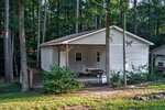 A small, white, single-story house with a covered front porch, picnic table, and ramp, surrounded by tall trees and greenery in a wooded area. Sunlight filters through the trees onto the grassy yard.