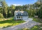 A two-story gray house with a white porch and attached garage sits on a large lot surrounded by tall trees and greenery, with a gravel driveway and a small shed in the backyard.
