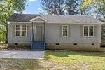 Single-story gray house with a small front porch and blue steps, two sets of double windows, and a grassy yard with a concrete driveway. Trees and blue sky are visible in the background.