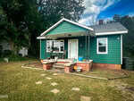 A small green house with white trim and a covered front porch, set on a grassy yard with stepping stones leading to the steps. There are potted plants and a rocking chair on the porch, with trees and a blue sky in the background.