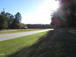 A rural, two-lane road curves through a grassy landscape bordered by trees. The sun is shining brightly, casting long shadows, and a truck is parked in the distance near the roadside.