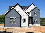 A newly constructed two-story house with a modern design features gray and white siding, multiple gables, and several uncurtained windows. The yard is unfinished with exposed soil and building materials nearby.