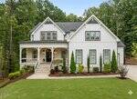 Two-story modern farmhouse with white board-and-batten siding, black roof, double front doors, covered porch, and neatly landscaped front yard with shrubs, trees, and a concrete driveway on the right.