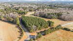 Aerial view of a rural landscape with a winding road passing through wooded areas and open fields, bordered by trees and patches of farmland under a partly cloudy sky.