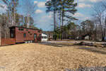 A modern tiny house on wheels sits in a cleared yard at 111-113 Red Oak Drive, Louisburg, surrounded by tall trees and neighboring homes under a partly cloudy sky. The ground is covered with straw and gravel.