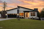 Modern single-story house with a dark roof, large black-framed windows, and a double garage. The lawn is neatly trimmed, and the sky is orange and pink at sunset. Trees surround the property.