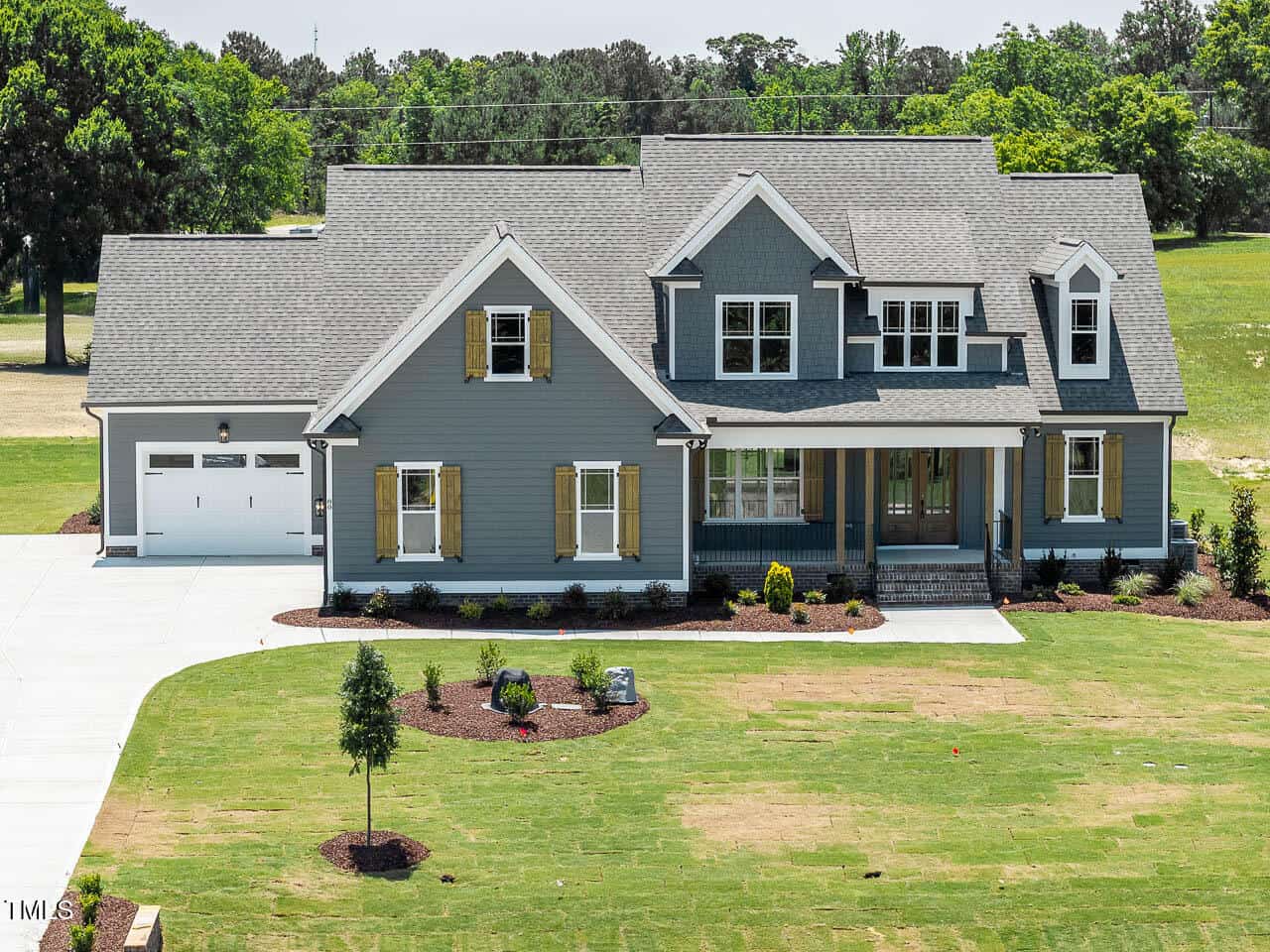 A two-story gray house with white trim, a covered front porch, dormer windows, and an attached two-car garage, surrounded by a partially landscaped yard with trees and grass.