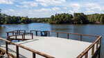 A wooden dock with railings extends over a calm lake, surrounded by trees under a partly cloudy sky. A blue slide is attached to the dock, and the water reflects the greenery and sky.