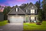 Two-story suburban house with gray siding, a white double garage door, black shutters, a covered front porch, manicured lawn, and trees in the background at sunset. Signs are displayed in the front yard of this charming Youngsville residence located at 510 Husketh Road.