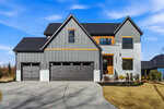 A modern two-story house with gray vertical siding, white brick, black-framed windows, and three dark garage doors. The home features natural wood accents and is surrounded by a newly landscaped yard.