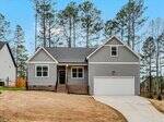 Single-story gray house with white trim, front porch with brick steps, and attached two-car garage. The yard has dry grass and is surrounded by tall pine trees under a clear sky.