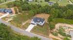 Aerial view of a two-story house with a gray roof and attached garage, located on a spacious lot with a driveway and surrounding grass, bordered by trees and neighboring homes.