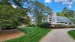 A white house with a steep roof and chimney sits beside a curved gravel driveway, surrounded by trimmed green grass, bushes, and tall pine trees under a partly cloudy sky.