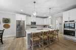 Modern kitchen with white cabinets, stainless steel appliances, a large island with a white countertop, and four wooden barstools. Pendant lights hang above the island, and wood flooring is throughout the space.