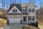 Two-story gray house with white trim, black shutters, a double garage, and a covered front porch. The yard is sloped and surrounded by leafless trees under a clear blue sky.
