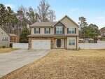 Two-story beige house with stone accents, green shutters, and a double garage. The home sits on a large lawn with a curved driveway and is bordered by a white fence and trees in the background.