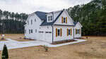 A modern two-story white house with black roof and shutters, wood accents, and attached garage, sits on a large, dry grassy lot bordered by pine trees under a cloudy sky.