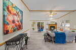 A living room with light-colored walls and floors, a ceiling fan, a beige sectional sofa with blue accents, a fireplace, large windows, and a colorful floral painting on the left wall.
