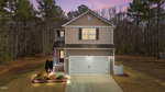 Two-story suburban house with beige siding, white trim, and a double garage. Lights are on inside and along the walkway. An American flag hangs near the front door. The house is surrounded by trees and lawn at dusk.