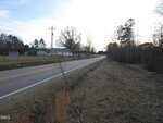 A two-lane road curves through a rural area, bordered by grassy shoulders and trees. Utility poles run alongside the road, and a few buildings are visible in the background under a partly cloudy sky.