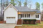 A white single-story house with a front porch, attached garage, brick foundation, and steps leading to the front door, surrounded by trees with no leaves and a concrete driveway.