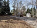 A gravel driveway at 112 Desoto Drive in Louisburg leads to two small sheds, one with a porch and the other with an American flag, surrounded by leafless trees and a grassy yard under a clear sky.