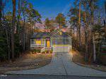 A gray house with a front porch, double garage, and lit windows sits among tall trees at sunset. A curved driveway leads from the street to the house, and the yard features simple landscaping.