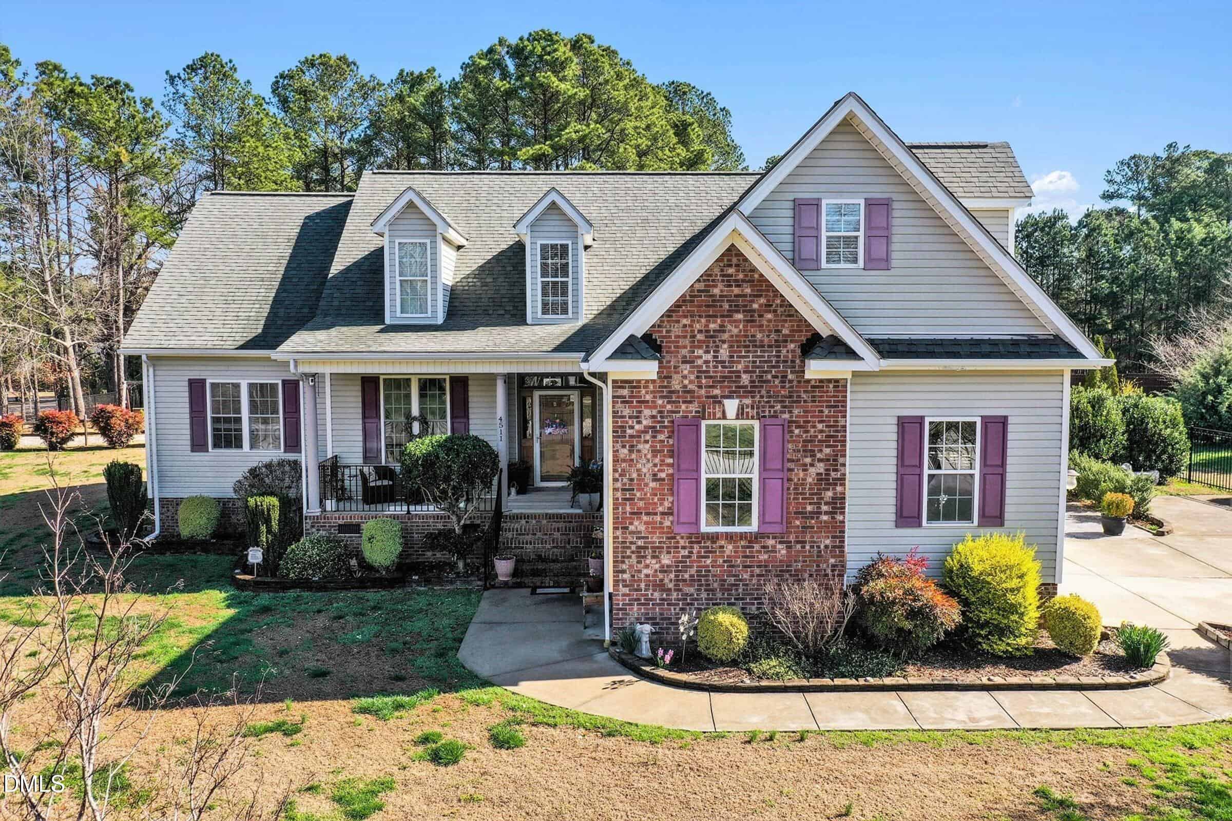 A two-story suburban house with beige siding, brick accents, and purple shutters, featuring a covered front porch, dormer windows, a neatly trimmed lawn, and a concrete driveway. Trees are visible in the background.