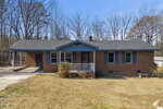 A single-story brick house with a dark roof, blue trim, and a small front porch sits on a patchy, dry lawn. Leafless trees fill the background under a clear sky. A driveway runs along the left side of the house.