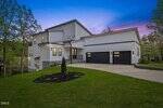 Modern two-story house with white siding, large black-framed windows, and a three-car garage. The home sits on a sloping lawn with landscaped mulch and young tree, set against a sunset sky and lush trees.