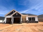 A single-story house under construction with white siding, dark gray gables, stone accents, and a covered front porch. The double garage is open, and the yard is unfinished with exposed dirt.