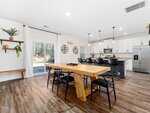 Modern open kitchen and dining area with wooden table, black chairs, island with barstools, stainless steel appliances, white cabinets, pendant lights, sliding glass doors, and wood flooring.