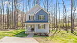 A two-story blue house with white trim sits in a grassy yard surrounded by tall, leafless trees. The house has a covered front porch, large windows, and a concrete driveway leading to a sidewalk.