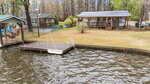 A small wooden cabin with a porch is situated on a grassy lot by the water, with a wooden dock and a small boat moored beside it; other cabins and trees are visible in the background.