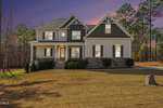 Two-story suburban house with gray siding, white trim, stone accents, and a covered front porch. The house sits on a large lawn with minimal landscaping, surrounded by tall trees at sunset.